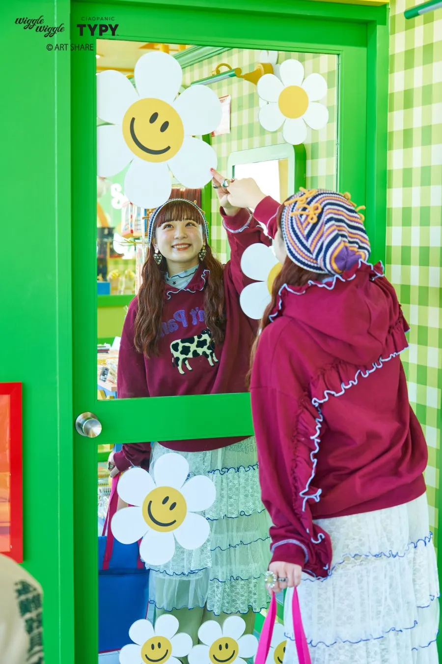 鏡に映る2人の女性。一人は赤い服を着ていて、もう一人は鏡に映っている。周りには花や緑の装飾が施されている。