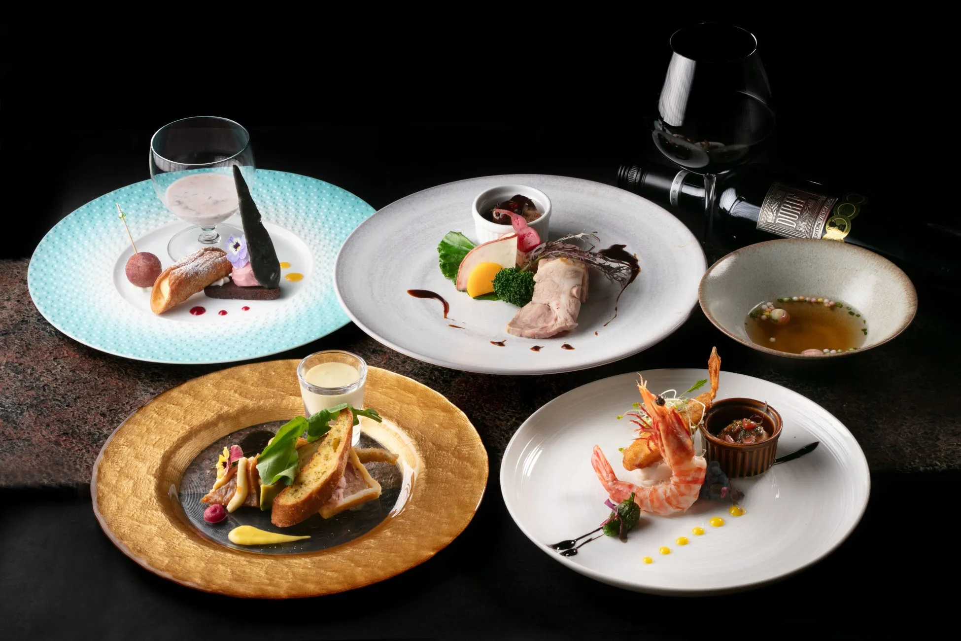 様々な料理が盛り付けられたコース料理の画像。デザート、スープ、前菜、メイン料理、エビ料理など、様々な料理が美しく配置されており、ワインボトルとワイングラスも添えられている
