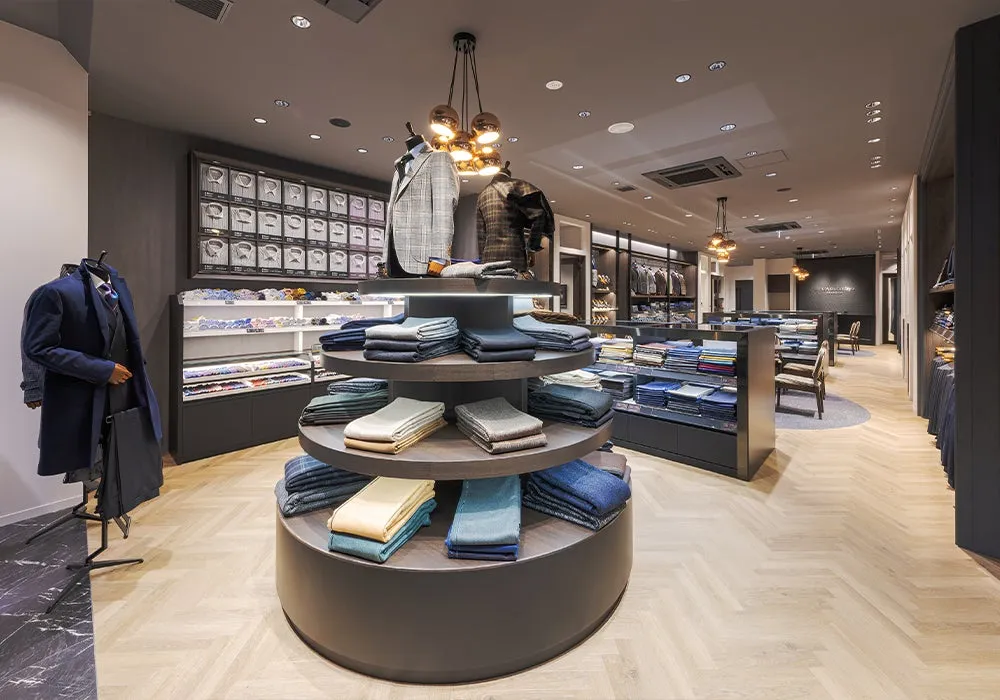 男性用スーツやシャツなどのアパレル商品が陳列された、洗練された雰囲気の店内。中央には円形のディスプレイがあり、色とりどりの生地が並べられています。奥には試着室とみられるスペースも見えます。