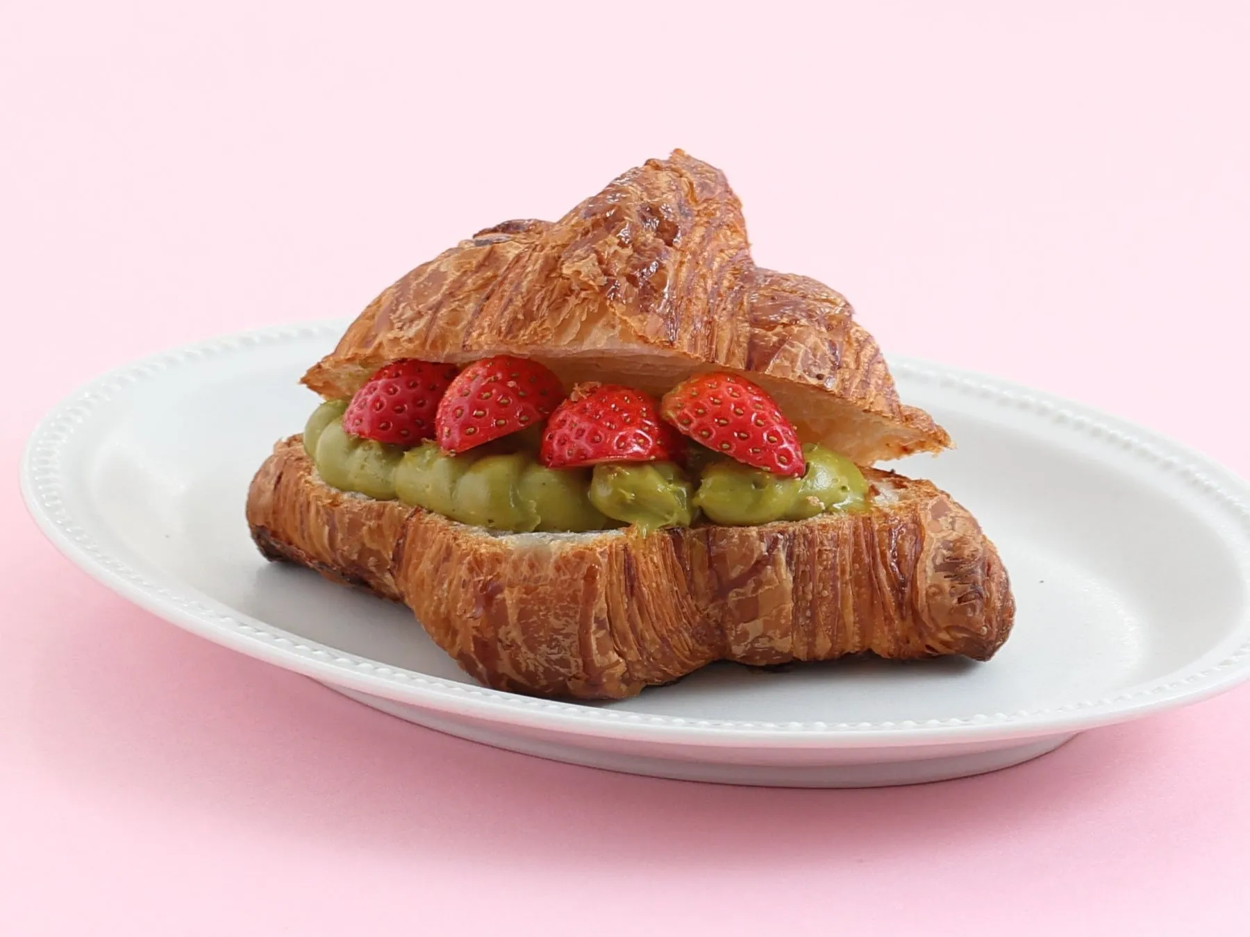 クロワッサンに緑色のクリームとイチゴが乗っている写真。白い皿の上に置かれ、背景はピンク色。