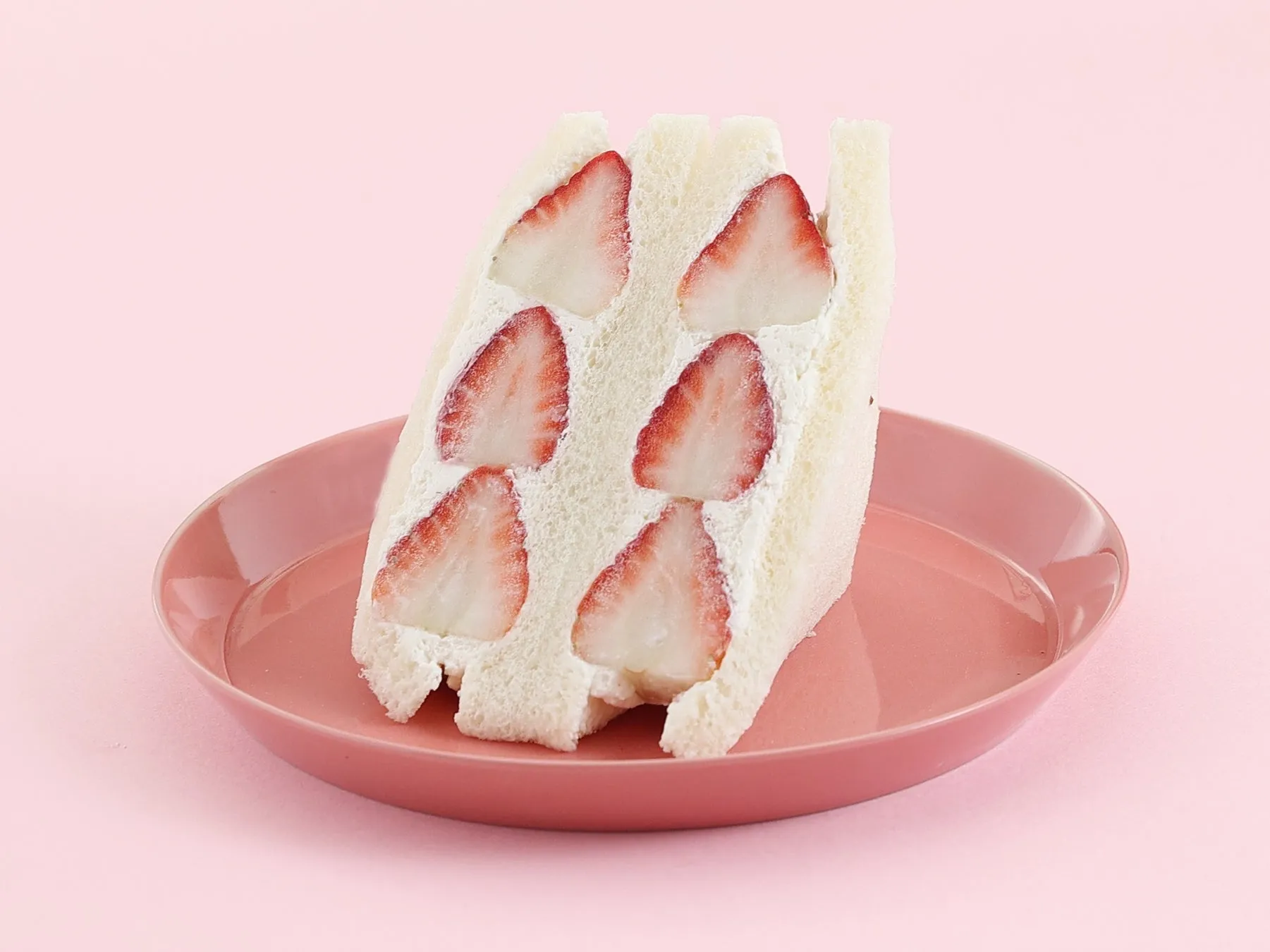 ピンク色の皿に載った、イチゴが挟まれたサンドイッチの写真です。サンドイッチは、白いパン、生クリーム、イチゴで構成されています。背景はピンク色です。