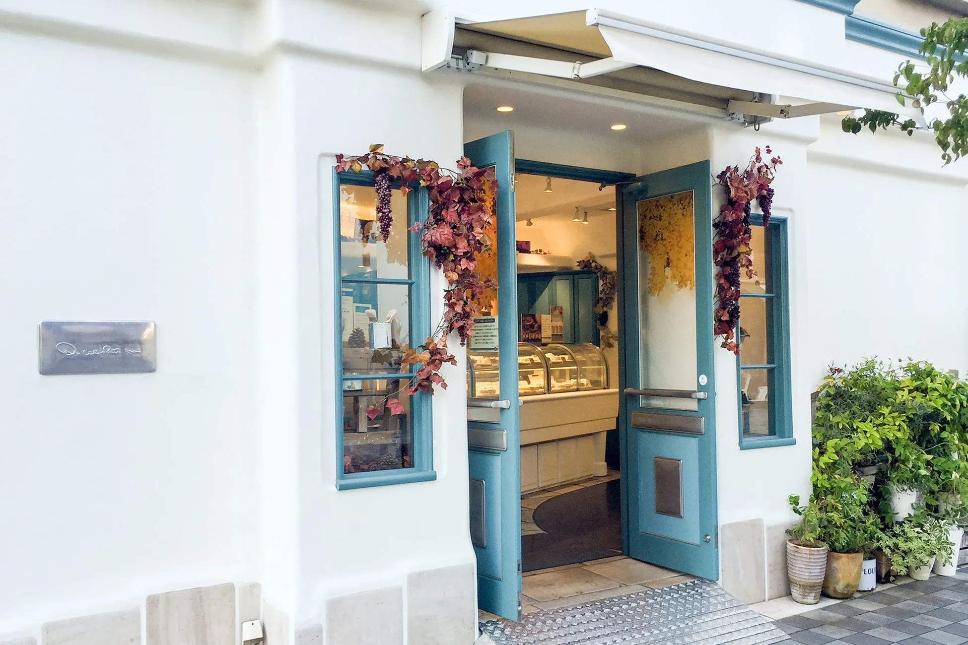 お店の入り口の写真。白い壁に、水色のドアと窓枠が印象的。ドアにはブドウの飾りがあり、店内にはケーキが並んでいる。