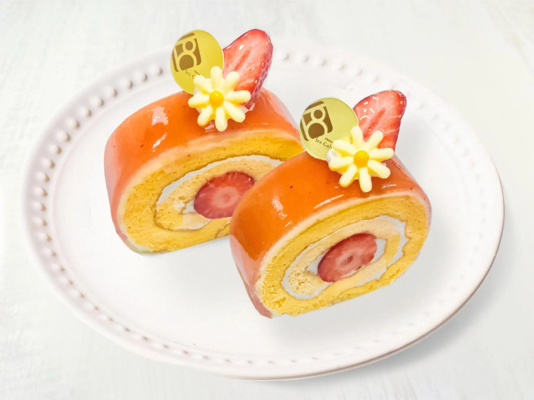 2つのロールケーキが白い皿に盛られている写真です。ロールケーキは、オレンジ色のゼリーで覆われ、中央にイチゴが配置されています。ロールケーキの上には、イチゴのスライスと黄色の花の飾りが施されています。皿は白い背景に置かれています。