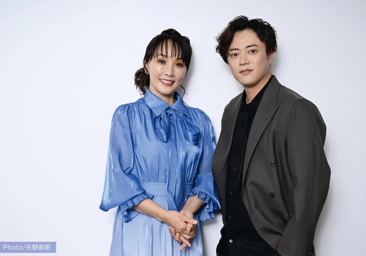 女性と男性が並んで立っている写真。女性は青いブラウスとスカートを着ており、男性はグレーのジャケットと黒いシャツを着ている。二人はカメラ目線で微笑んでいる。