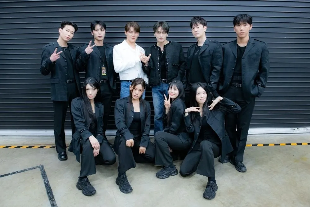 9人のグループがポーズをとって写真撮影をしている様子。一部のメンバーは黒いスーツを着ており、他のメンバーは白いシャツを着ている。