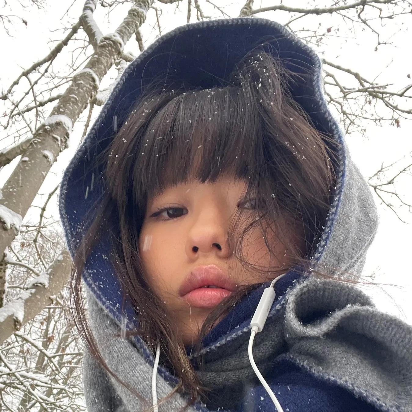 雪の中でフードを被った女性のクローズアップ写真。イヤホンをしており、周りには雪が降っている。