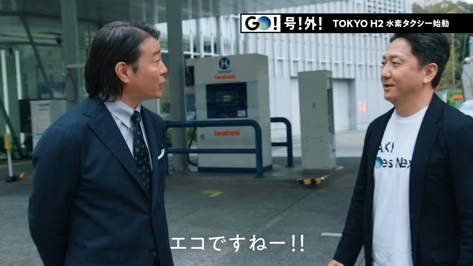水素ステーションの前で話す2人の男性。1人はスーツ姿、もう1人はTシャツにジャケット姿。背景には「TOKYO H2 水素タクシー始動」の文字。