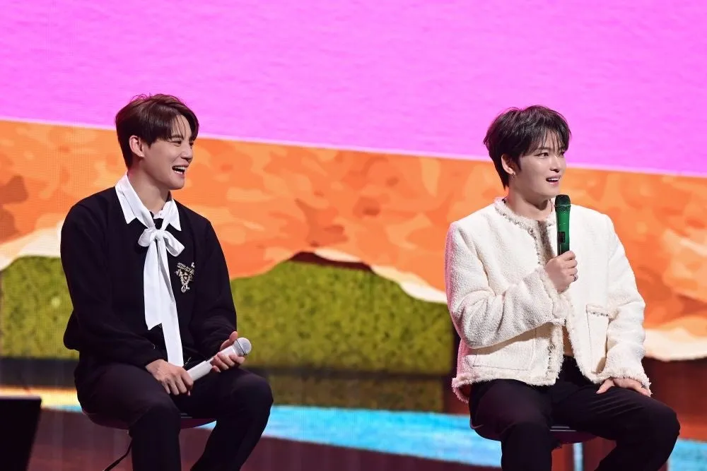 2人の男性が座ってマイクを持っている写真。背景はピンク色とオレンジ色で、ステージのようだ。