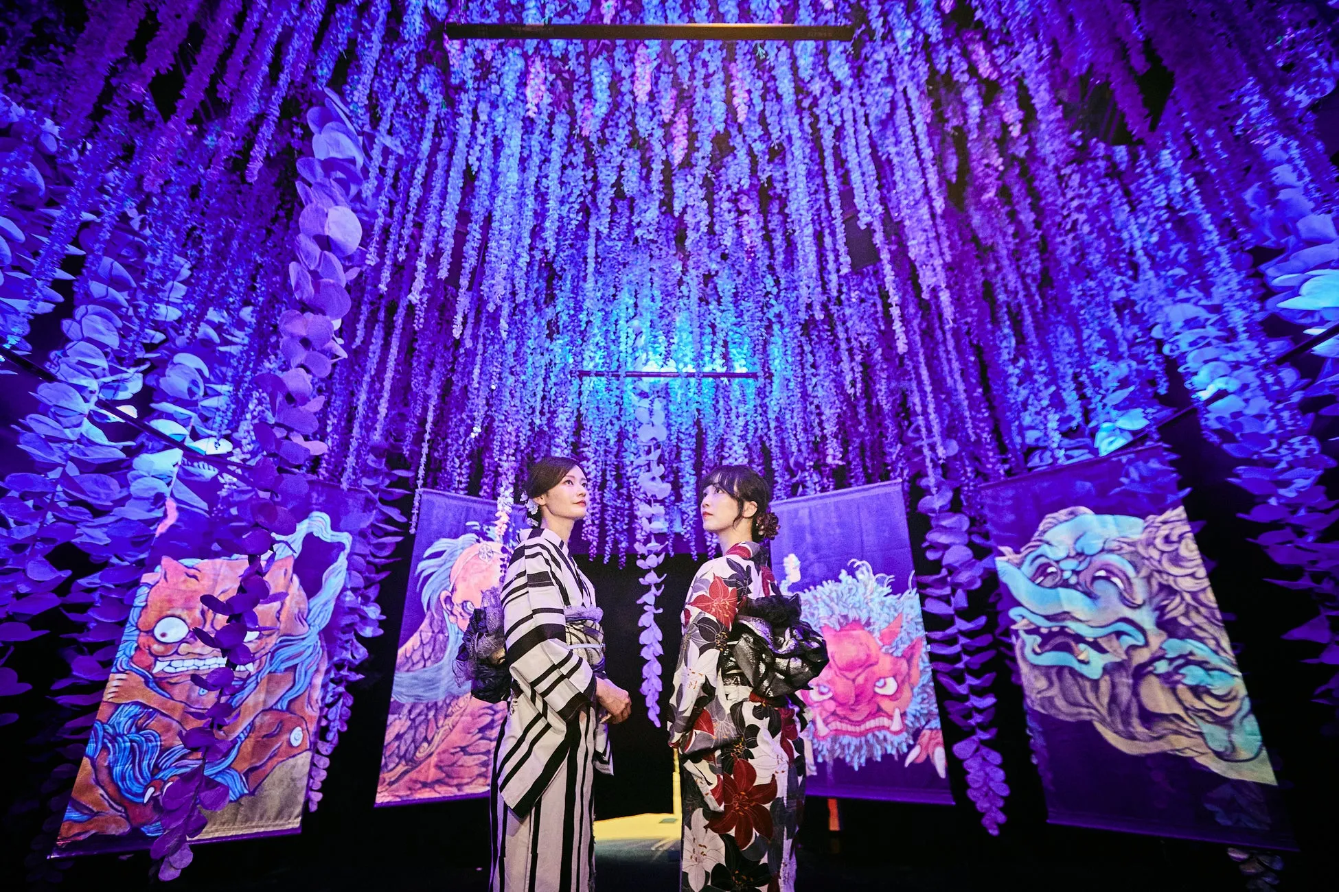 2人の女性が着物を着て、紫色の装飾が施された空間に立っています。背景には、日本の伝統的な絵画が飾られています。