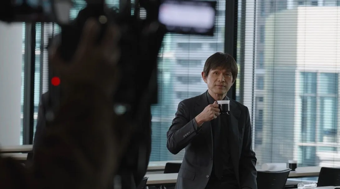 コーヒーカップを手にした男性がカメラの前で立っている写真です。彼は黒いスーツを着ており、窓のそばに立っています。