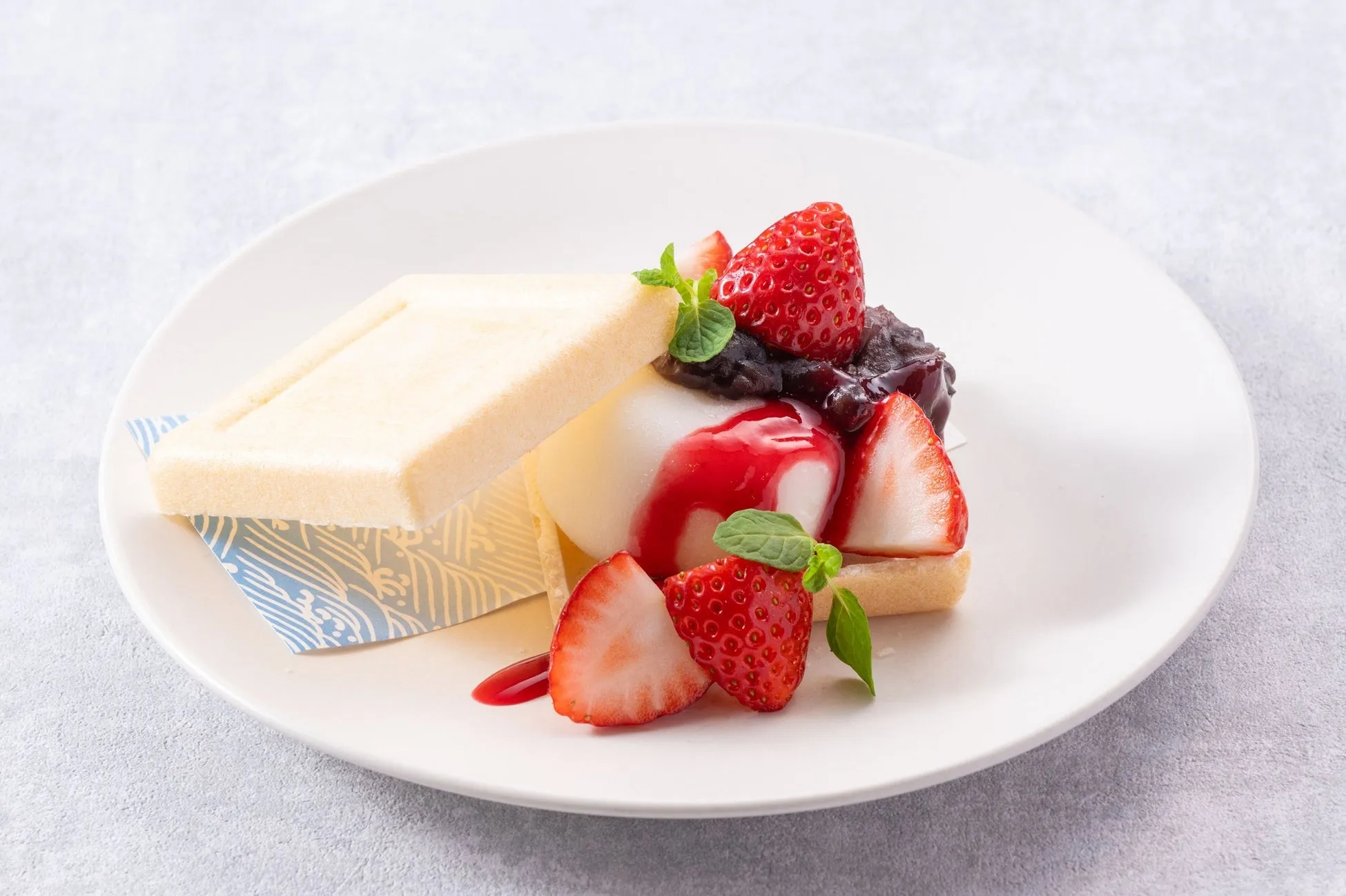 白い皿の上に、イチゴ、あんこ、ソース、ミントでデコレーションされたデザートが乗っています。デザートは、クッキーのようなものと、白いスポンジのようなものに挟まれています。