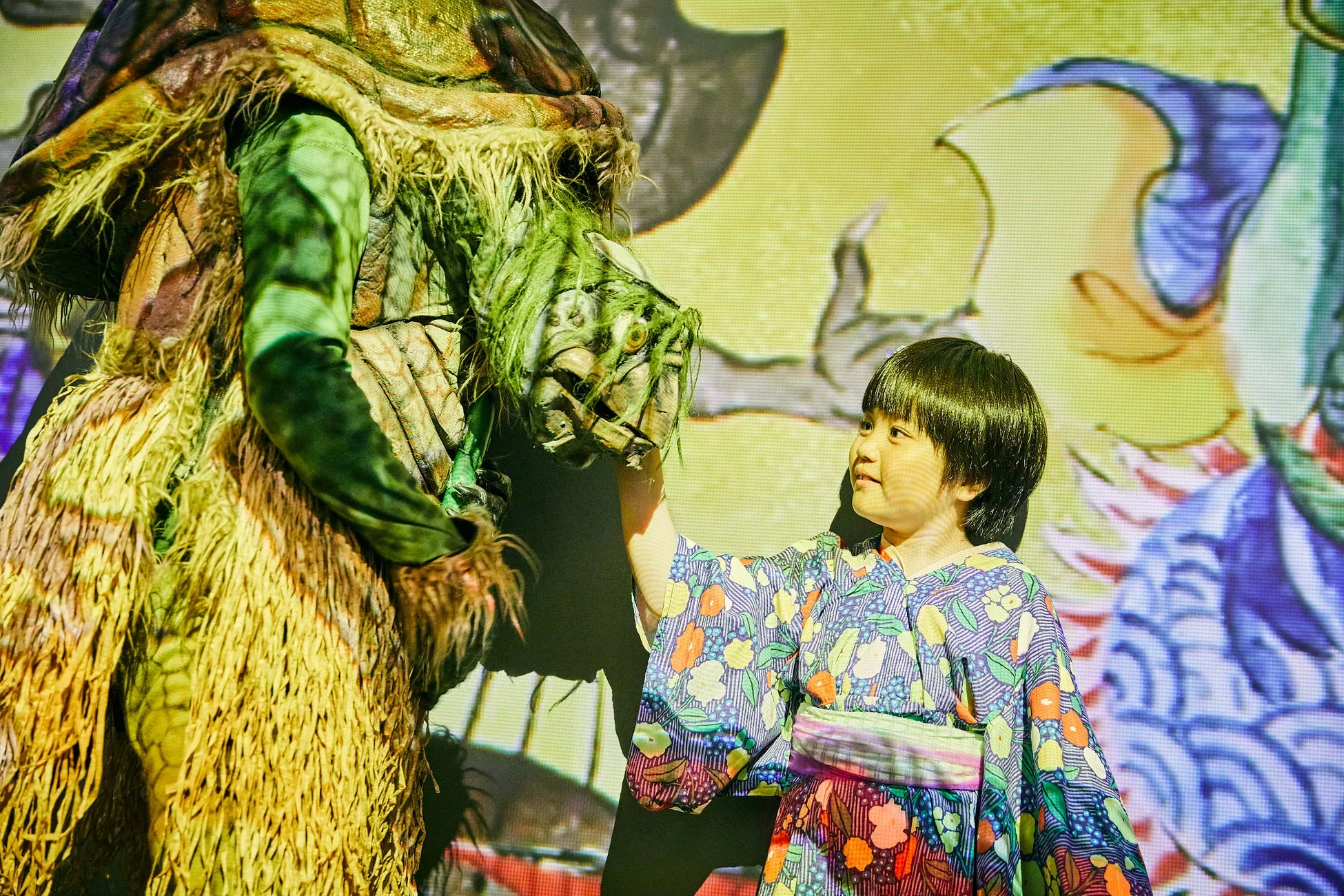 子供が、着ぐるみのキャラクターに触れている様子を捉えた写真です。子供はカラフルな浴衣を着ており、キャラクターは緑色の草のようなもので覆われています。背景には、イラストが映し出されています。
