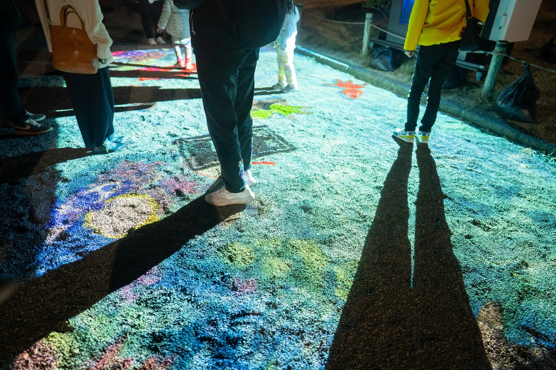 カラフルな光が地面に投影された、夜間の屋外シーン。人々がその光の中を歩いている。