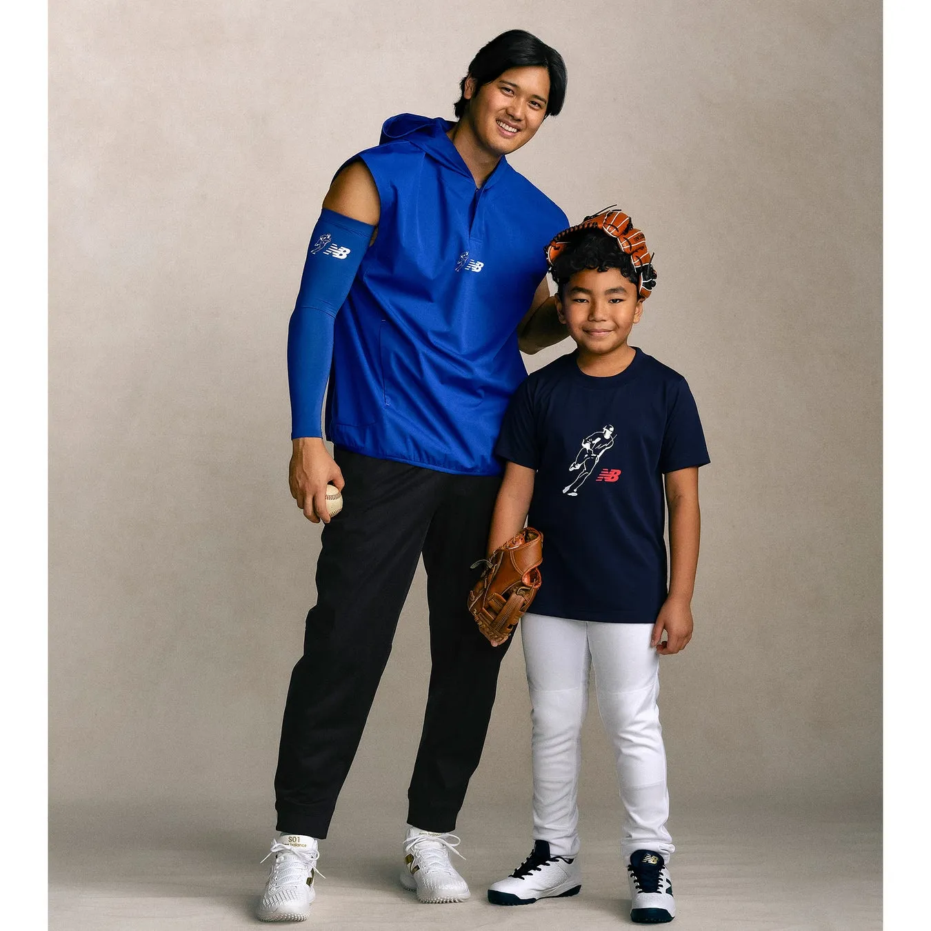 男性と子供が野球のユニフォームを着て、笑顔で立っている写真。男性は青いジャケットと黒いパンツ、白いスニーカーを履き、野球ボールを持っています。子供は紺色のTシャツと白いパンツ、野球グローブと帽子、白いスニーカーを履いています。