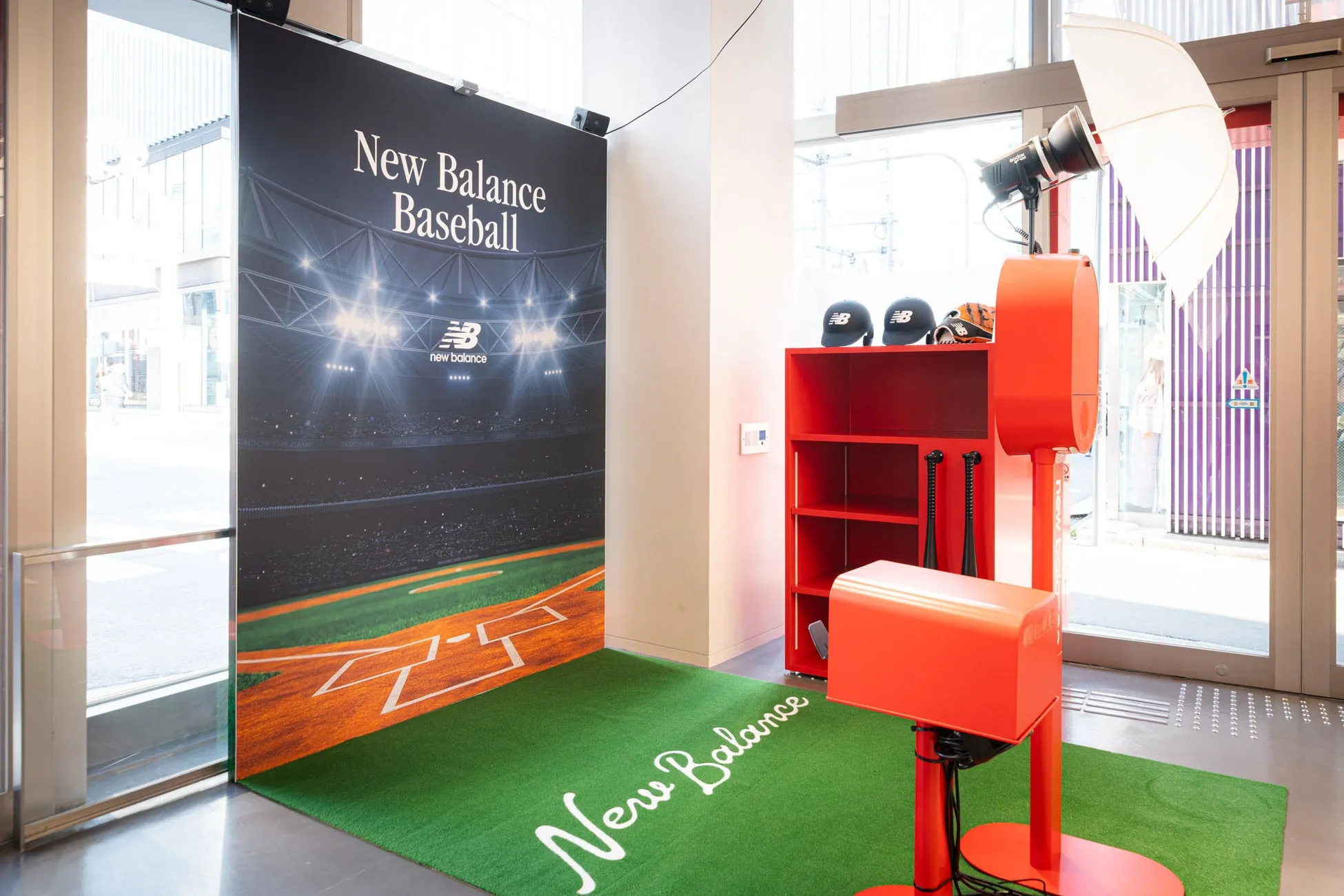ニューバランスの野球関連イベントスペースの写真。背景には野球場のグラフィックがあり、緑の芝生には「New Balance」のロゴが描かれています。赤い棚には野球帽やグローブが置かれ、写真撮影用の照明器具も設置されています。