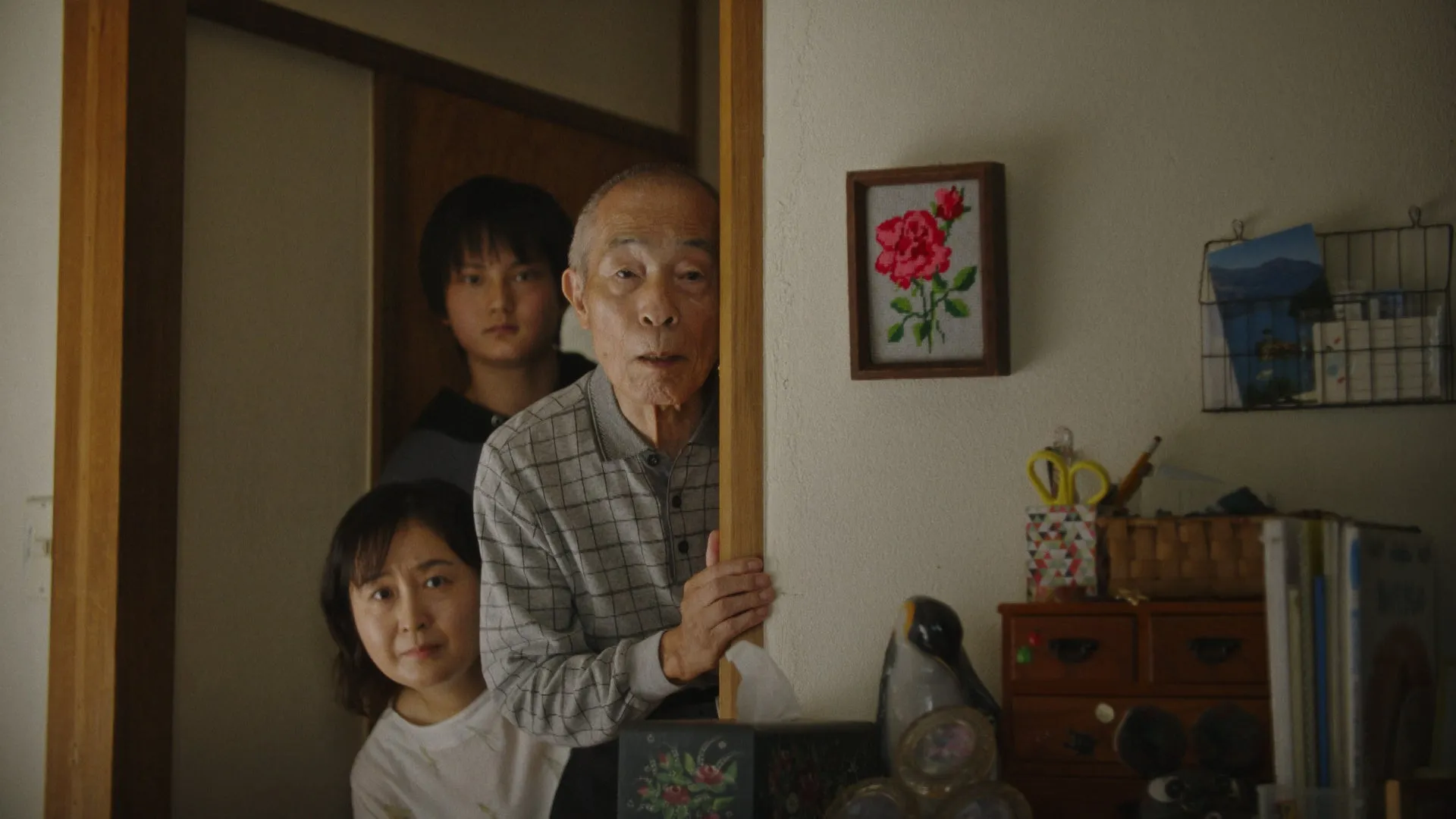 ドアの隙間から顔を覗かせる3人の人物。手前には高齢男性、その後ろに若い男性と女性がいます。