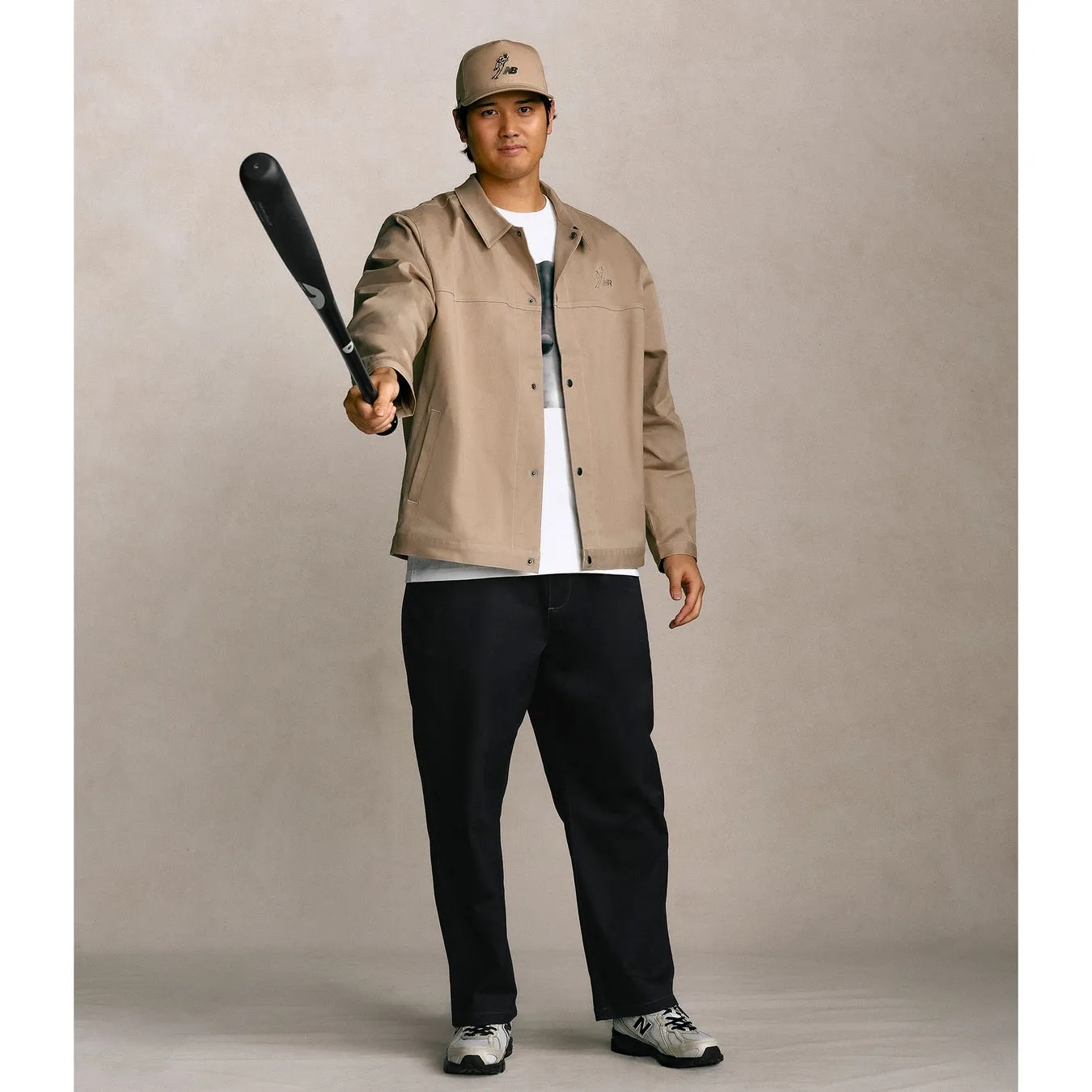 野球のバットを持った男性の写真。彼はベージュのジャケット、黒いパンツ、帽子をかぶっており、背景は単色です。