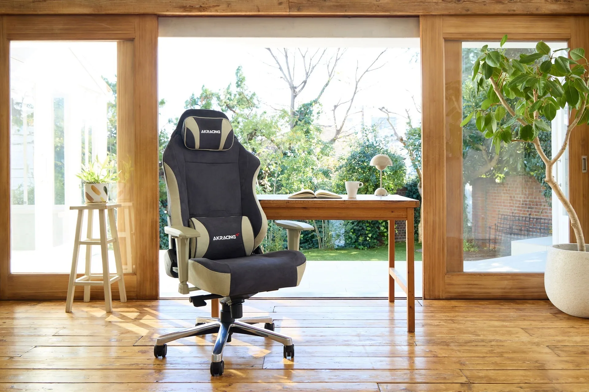 ゲーミングチェアが置かれた明るい部屋の様子。木製の窓から庭の緑が見え、テーブルの上には本やカップ、照明が置かれている。床は木製で、植物が配置され、リラックスした雰囲気。