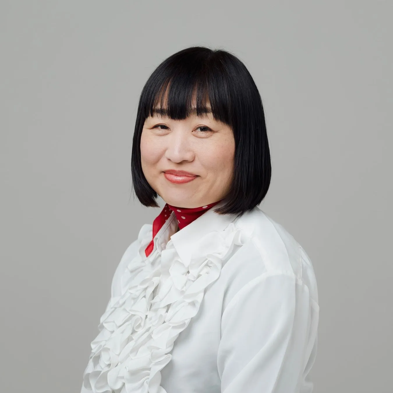 白いフリルシャツを着て、赤いスカーフを首に巻いた、黒髪の女性のポートレート。彼女はカメラに向かって微笑んでいます。