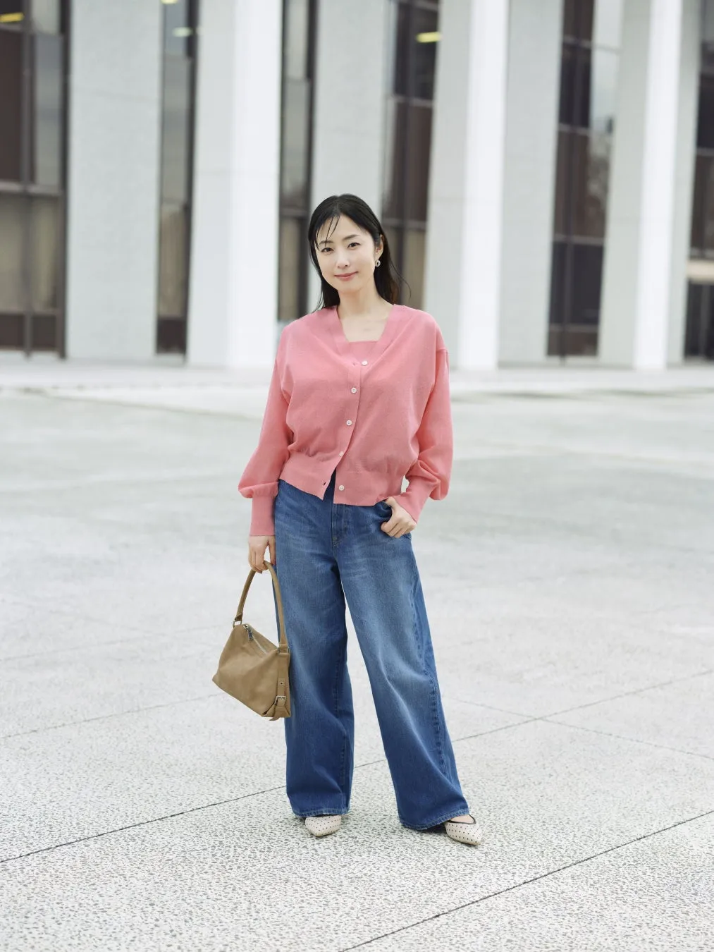 ピンクのカーディガンとジーンズを着た女性が、ベージュのバッグを持って立っている写真です。背景には、白い建物があります。