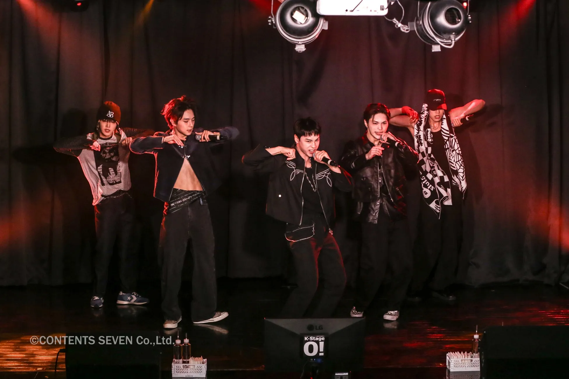 5人の男性が舞台でパフォーマンスをしています。彼らはカジュアルな服装をしており、マイクを持っていたり、ダンスのポーズをとっています。背景は暗く、照明が当たっています。