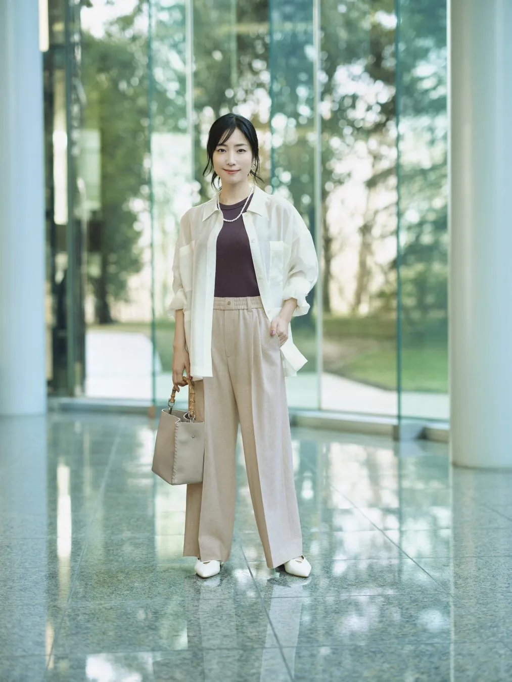 ガラス張りの建物の前に立つ女性。白いシャツ、茶色のトップス、ベージュのパンツ、白い靴を履き、ベージュのハンドバッグを持っている。彼女は穏やかな表情でカメラを見つめている。