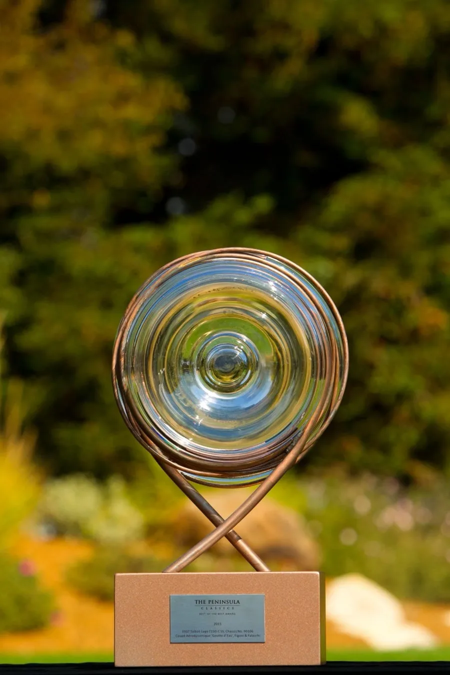 屋外の緑豊かな背景に、透明なガラスと銅色の金属コイルが特徴的なデザインの「THE PENINSULA CLASSICS BEST OF THE BEST AWARD 2015」のトロフィーが写っています。