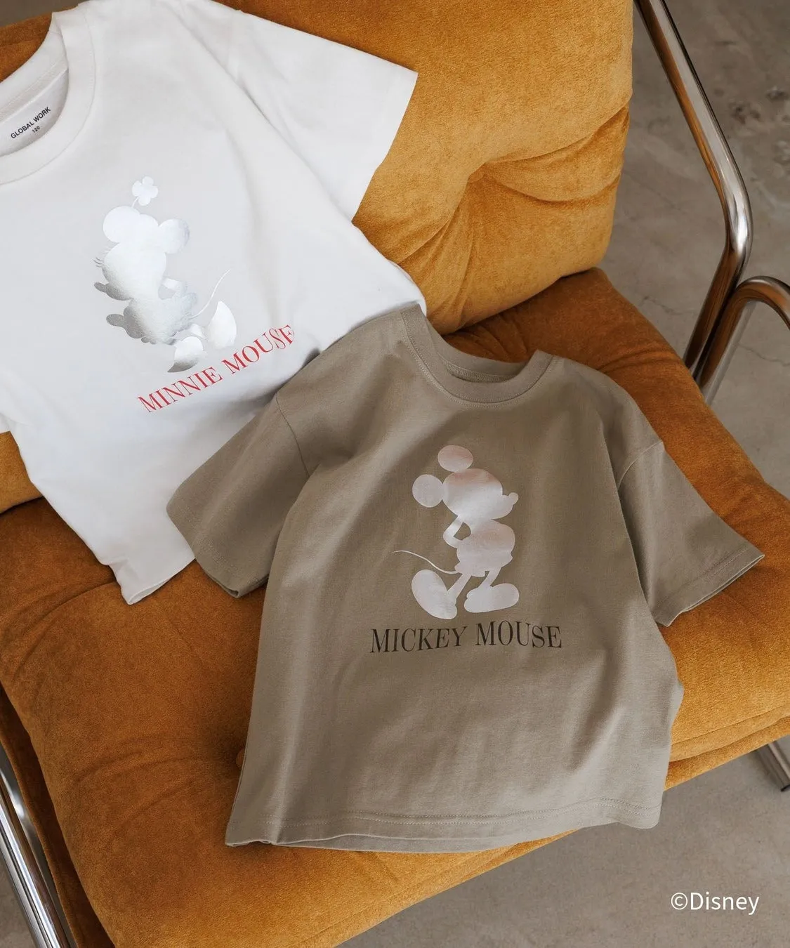 ミッキーマウスとミニーマウスのプリントがされたTシャツが2枚、オレンジ色の椅子の上に置かれている様子を写した写真です。白いTシャツにはミニーマウス、茶色のTシャツにはミッキーマウスがプリントされています。