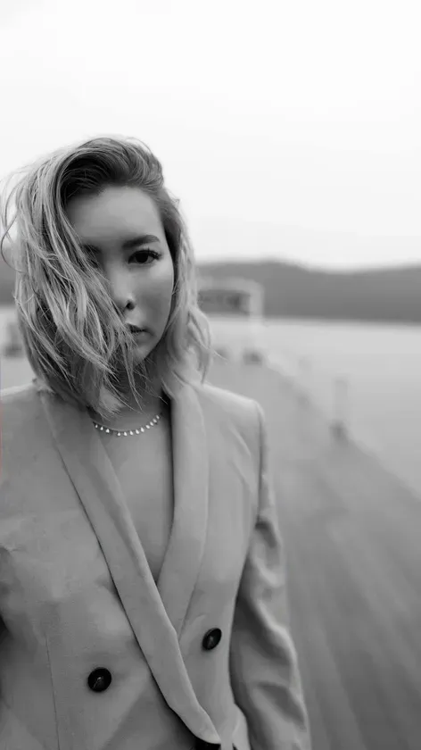 女性がポートレート写真でポーズをとっています。彼女はジャケットを着ており、カメラを見ています。背景には水辺とぼやけた建物があります。