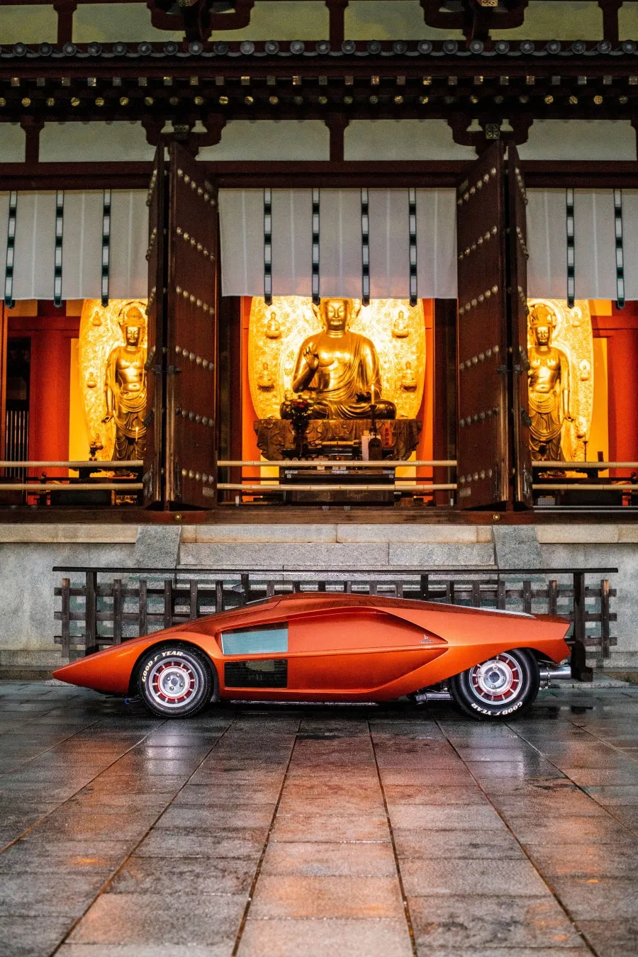 日本の伝統的な寺院を背景に、未来的なオレンジ色のコンセプトカーが展示されている様子。金色の仏像が並ぶ厳かな空間と、流線型の車体が対照的で、異文化が融合した独特の雰囲気を持つ一枚です。
