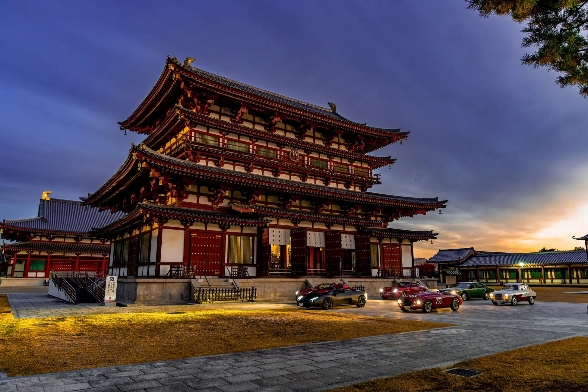 夕暮れ時か夜明けの美しい空の下、日本の伝統的な寺院がライトアップされ、その前に数台のクラシックカーやスポーツカーが並べられています。古き良き建築と現代的な車の対比が印象的な一枚です。