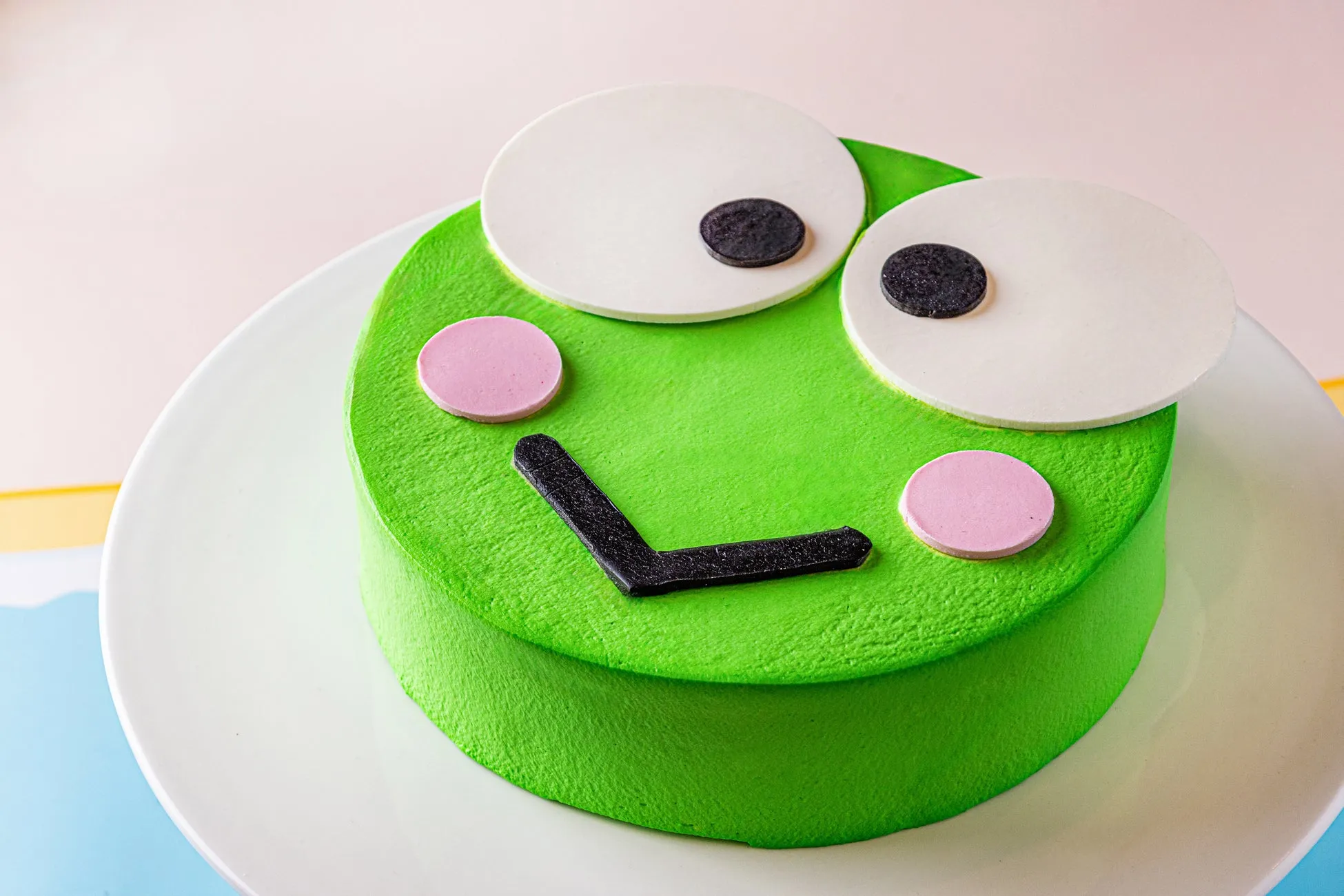 カエルの顔を模したケーキが白い皿の上に置かれています。ケーキは緑色で、白い丸い目、黒い瞳、ピンクの頬、黒い口が特徴です。