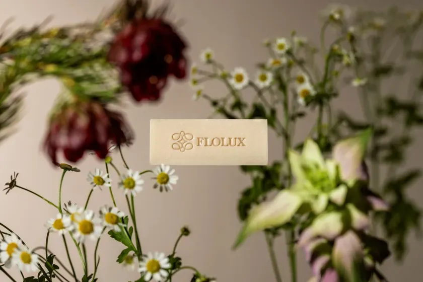 石鹸と花が写った画像です。石鹸には「FLOLUX」の文字が刻まれています。背景にはカモミールのような白い花や、赤い花が見えます。