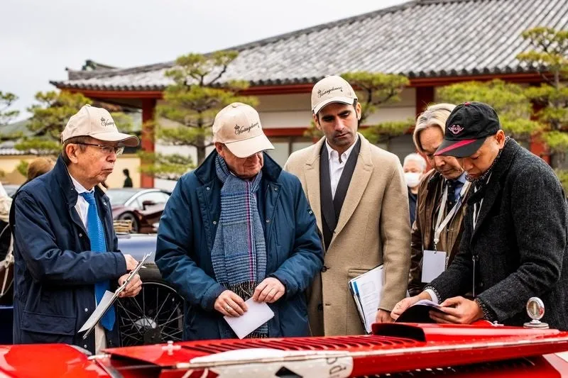 複数の男性が屋外で赤いヴィンテージカーを囲み、興味深そうに観察したり議論したりしている様子です。背景には日本風の建物が見え、何らかのイベントが開催されているようです。