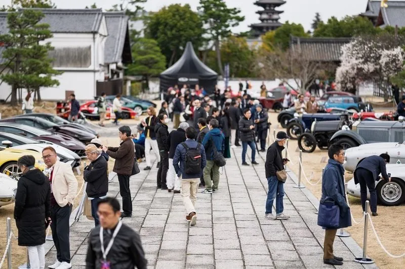 多くの来場者が、現代のスポーツカーからクラシックカーまで多様な自動車が展示された屋外イベントを楽しんでいる様子。日本の伝統的な建築物を背景に、人々が車両を鑑賞している。