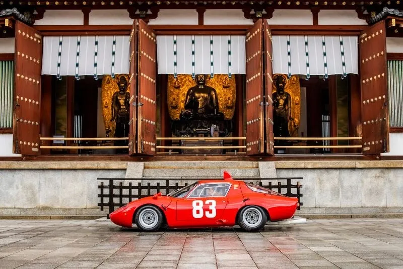 日本の伝統的な寺院を背景に、真っ赤なクラシックカー「83」が展示されている様子を捉えた画像です。歴史と現代の融合が印象的です。