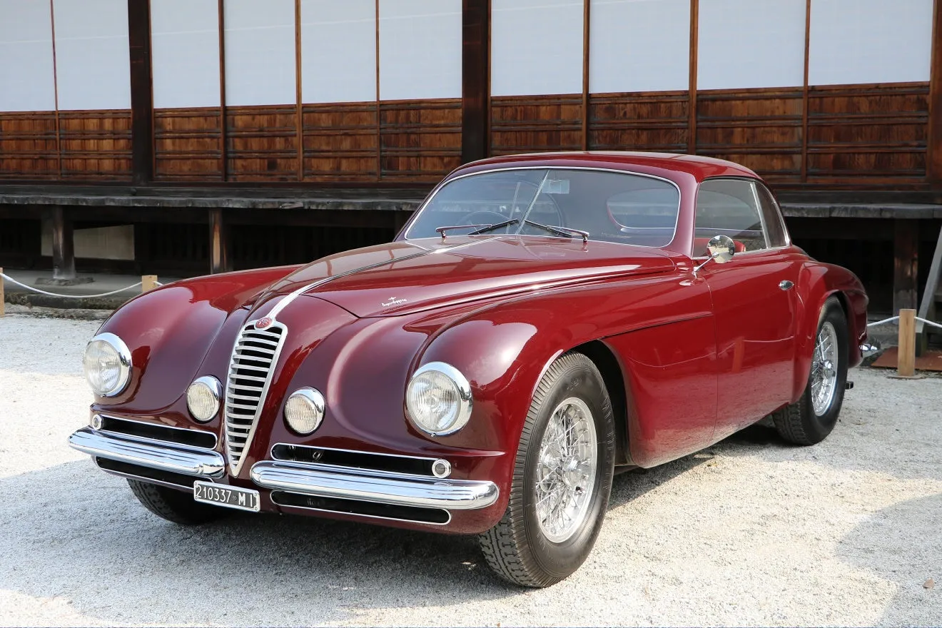 伝統的な日本家屋を背景に、美しいワインレッドのクラシックカーが展示されている。この車は優雅な曲線を持つヴィンテージのアルファロメオで、そのレトロなデザインが際立っている。